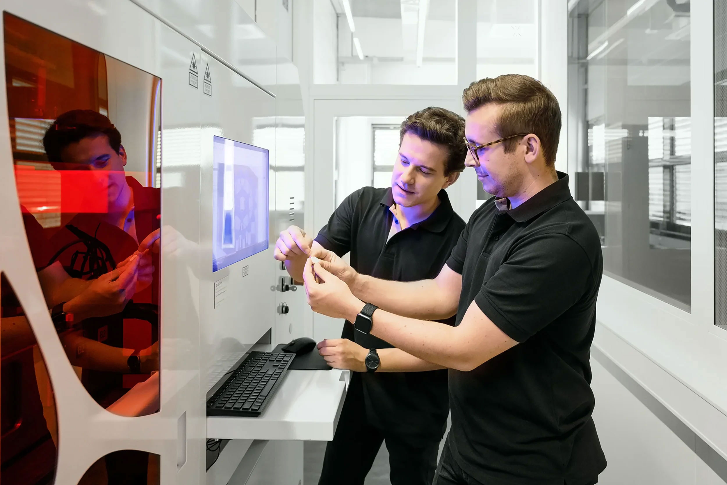 Two men in black polo shirts examining a small 3d printed object together in a modern lab with white walls and a computer workstation.