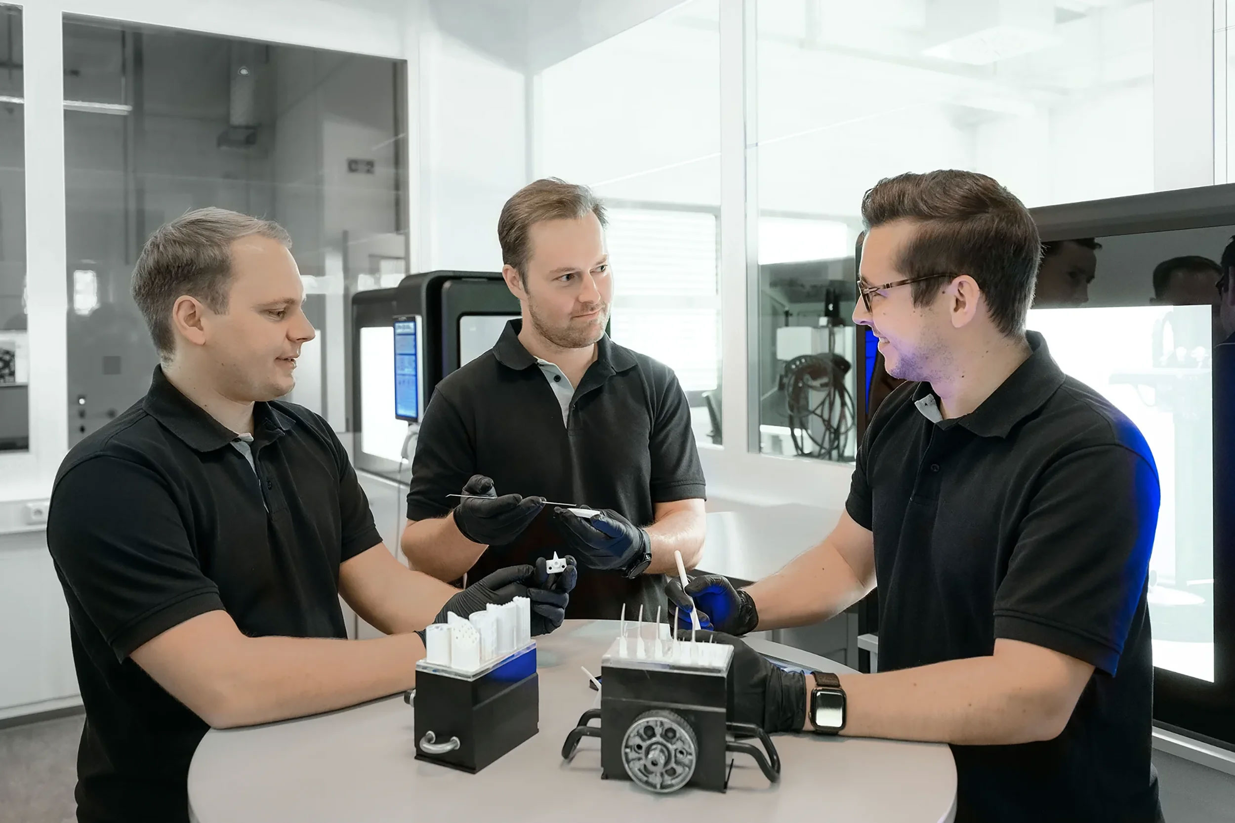 Three men in black polo shirts and gloves discussing and examining small white 3D printed objects on a table in a modern lab setting.