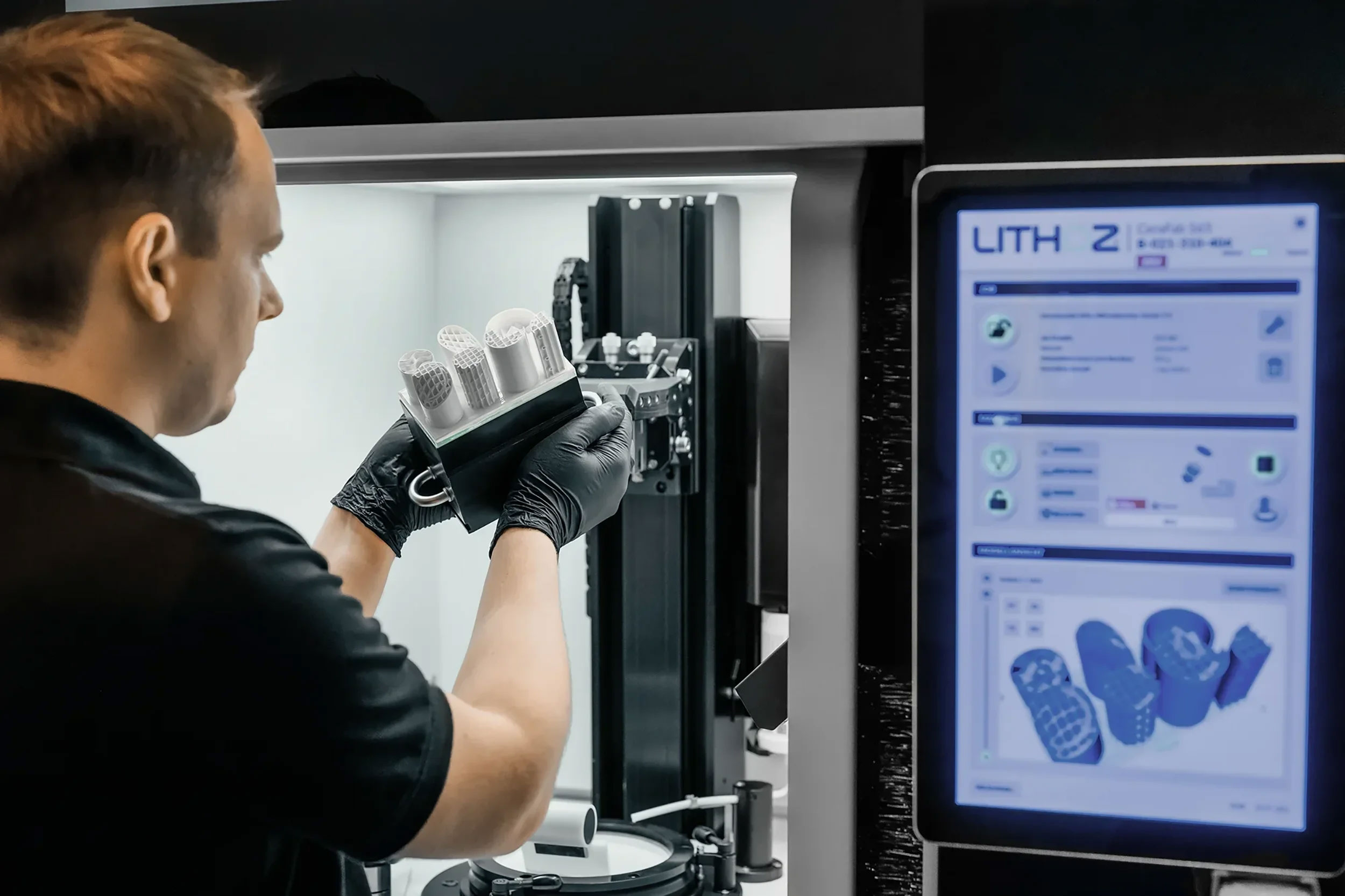 Man wearing black gloves inspecting a 3D-printed object inside a 3D printing machine with a touchscreen display showing the print design.