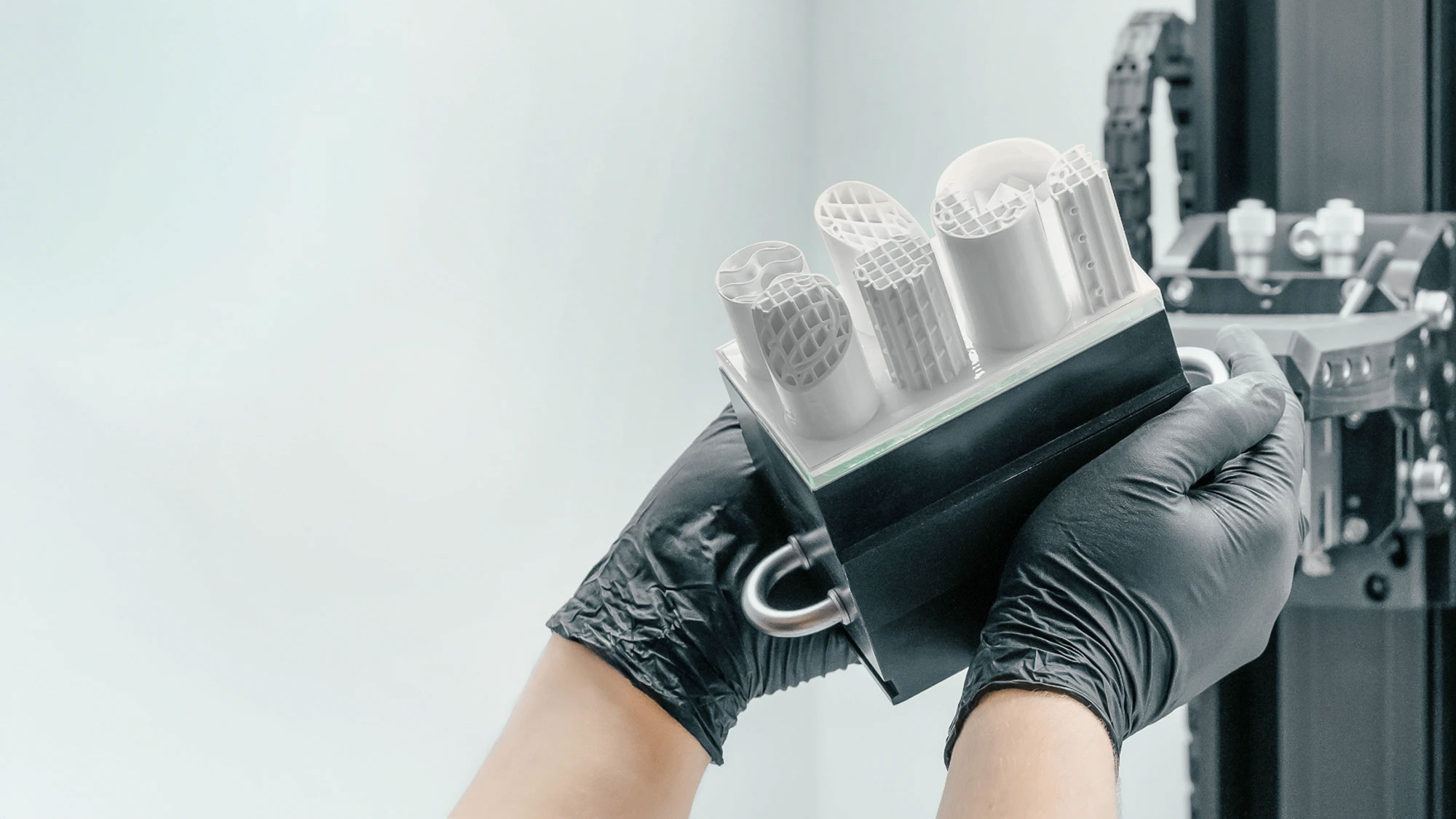 Person wearing black gloves holding a black device with five white cylindrical samples of ceramic 3d printing with internal lattice structures.