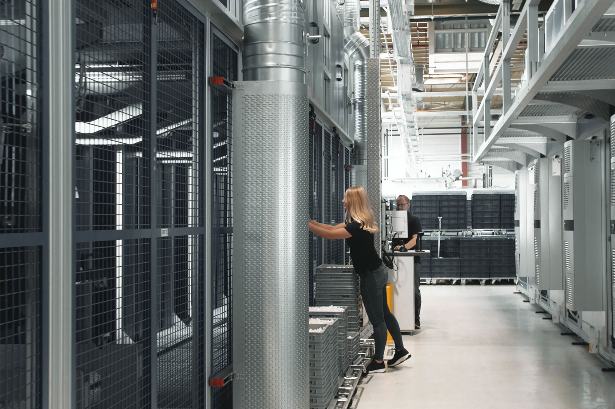 A woman operating a machine in a modern industrial facility with another worker at a computer station in the background.