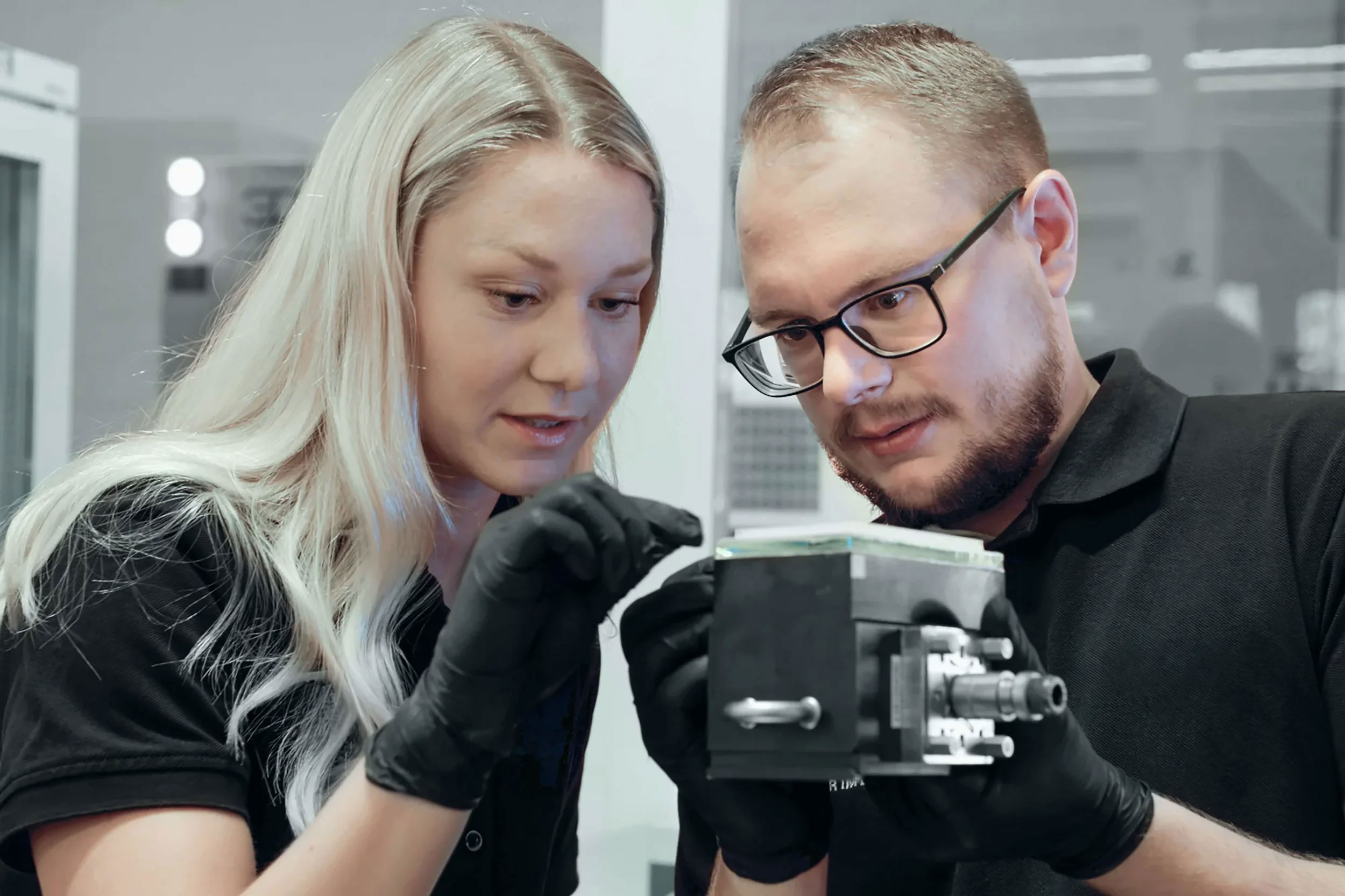 Two engineers wearing black gloves and black shirts closely inspecting a 3d printer part in a lab setting.