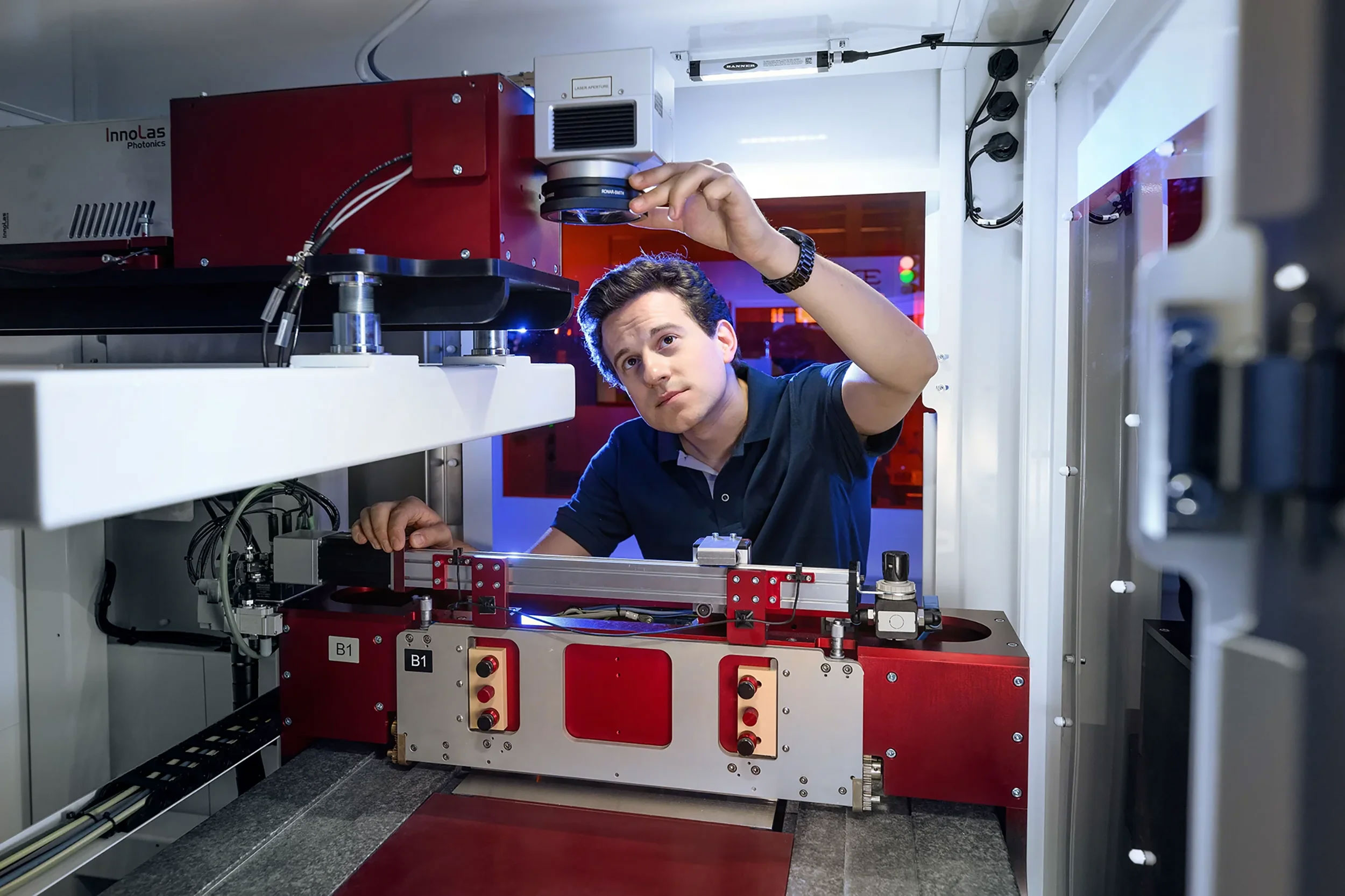 Technician inspecting a red and white 3d printing machine inside a lab or industrial setting.