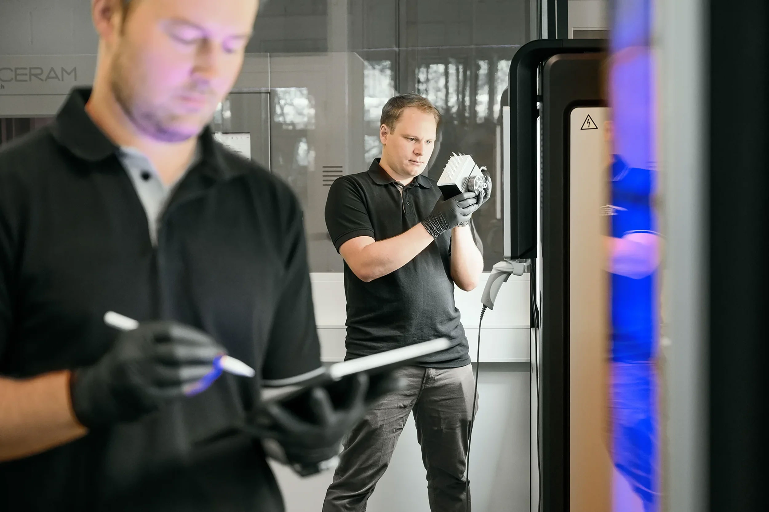 Two men in black shirts and gloves working in a lab, one holding a tablet and stylus, the other inspecting a 3d ceramic printer.