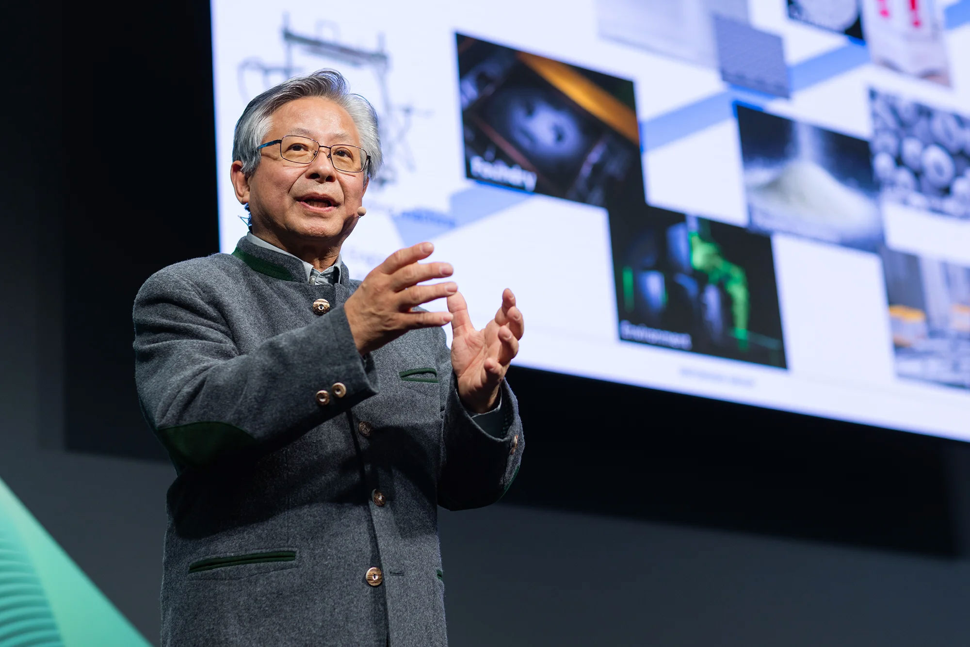 man speaking and gesturing with hands in front of a large presentation screen.