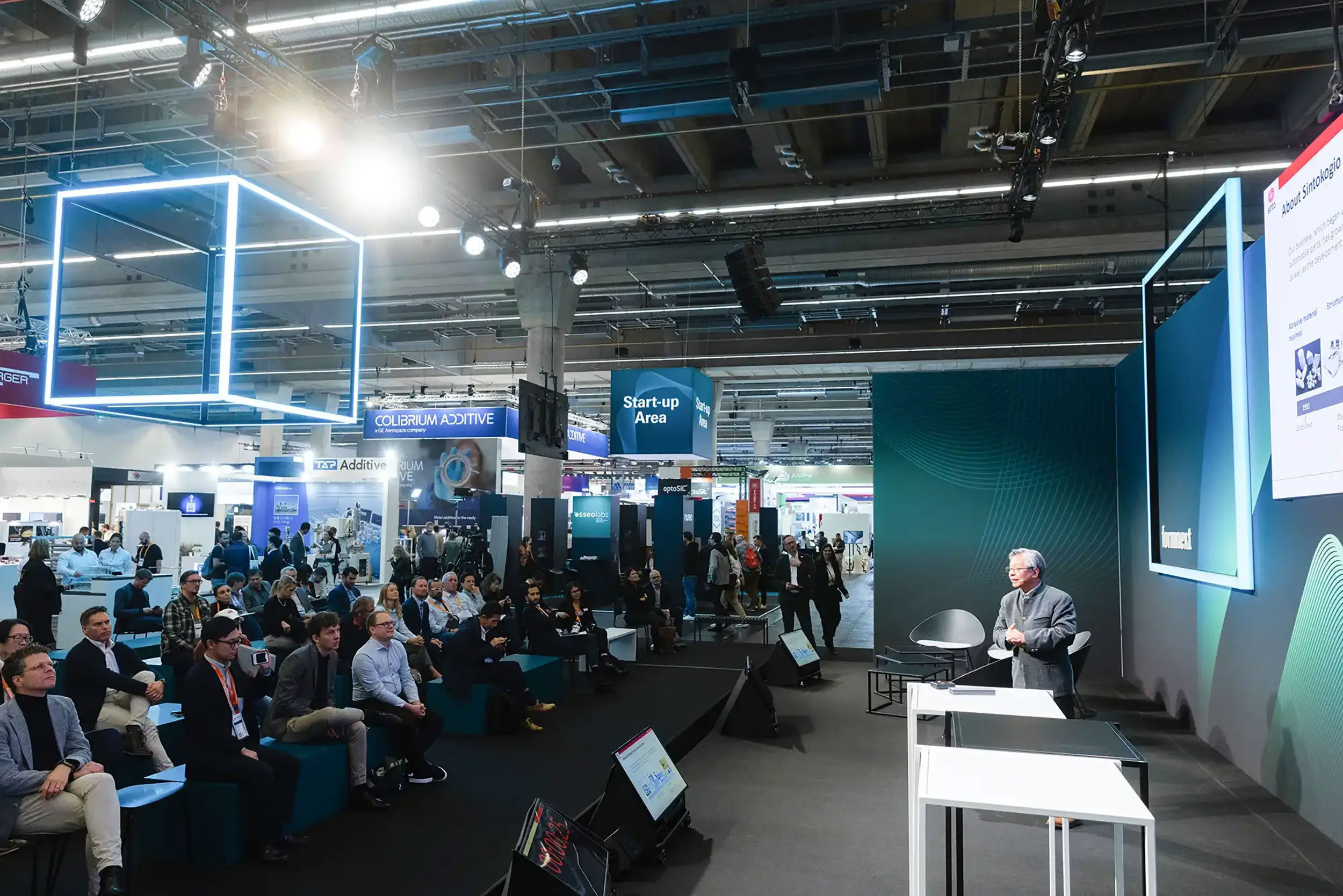 Speaker presenting on stage to an audience seated at a conference or trade show with illuminated booths in the background.